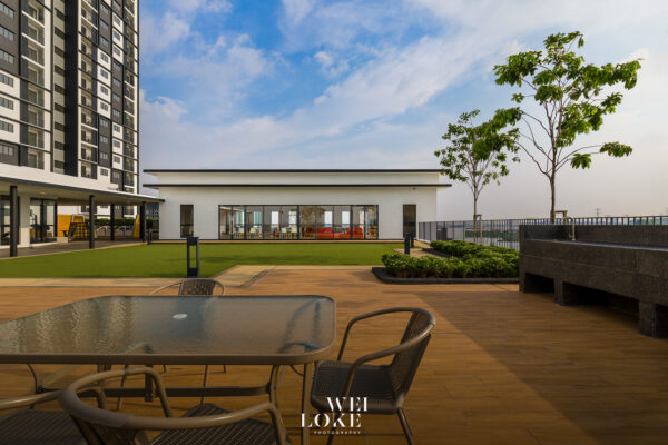 Modern apartment complex courtyard with poolside seating, a white glass-walled clubhouse, and a tall residential tower on the left under a blue sky.